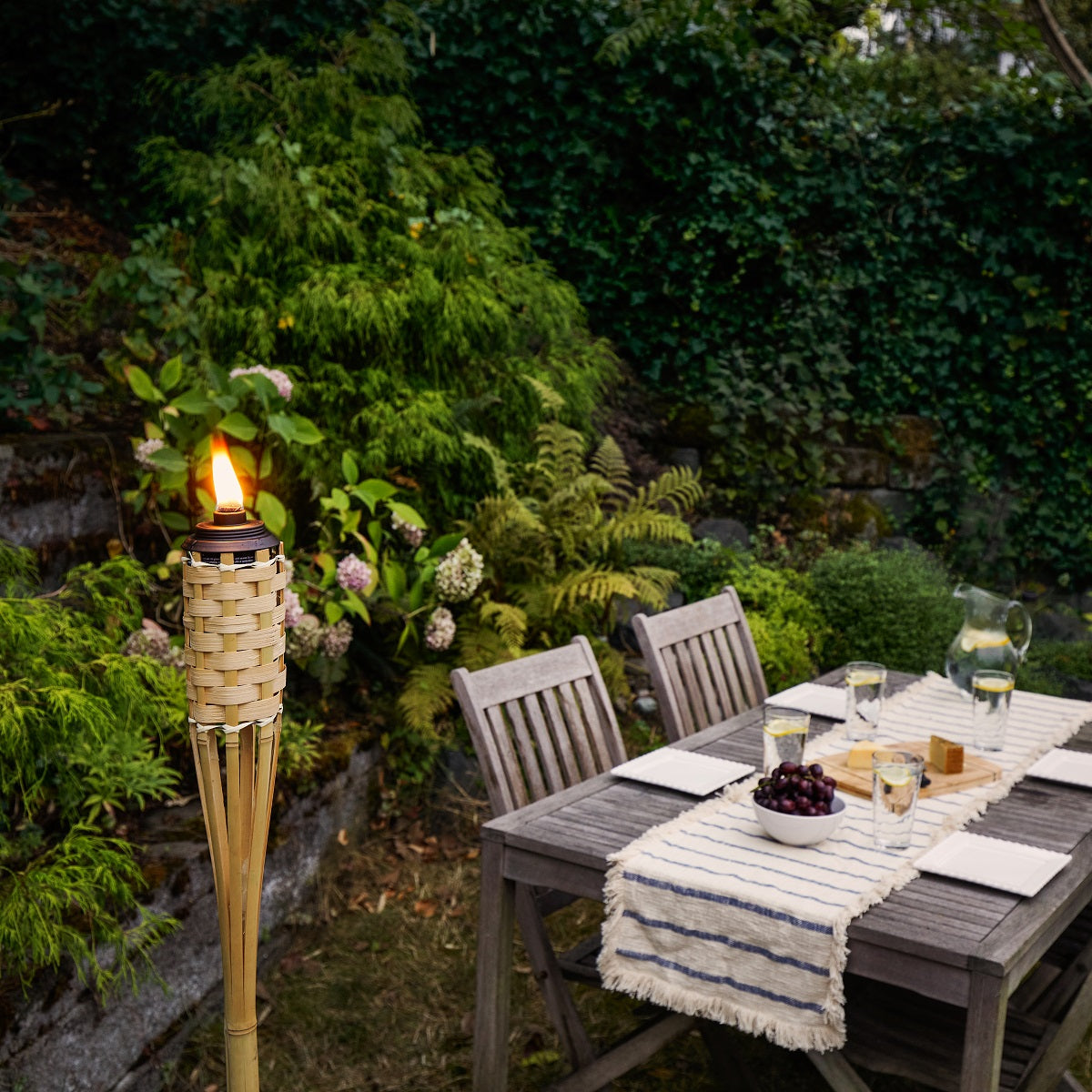 Lit bamboo TIKI® torch beside an outdoor dining table set for four in a lush, green backyard garden.