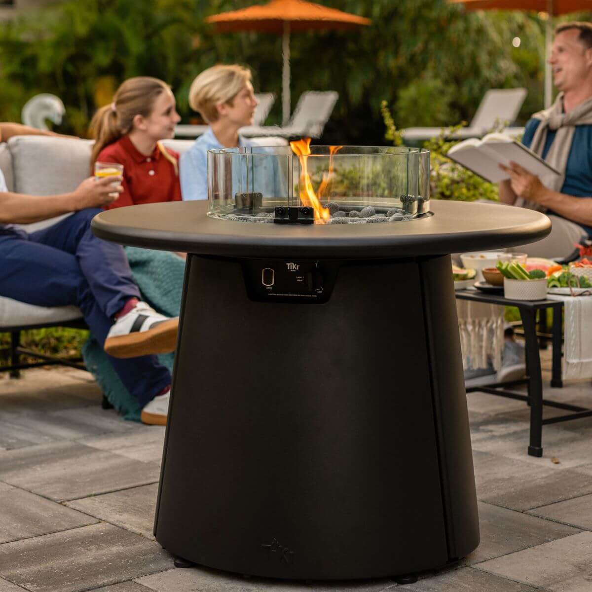 Modern TIKI® fire pit table with glass guard, surrounded by people relaxing outdoors on a patio with food and drinks.