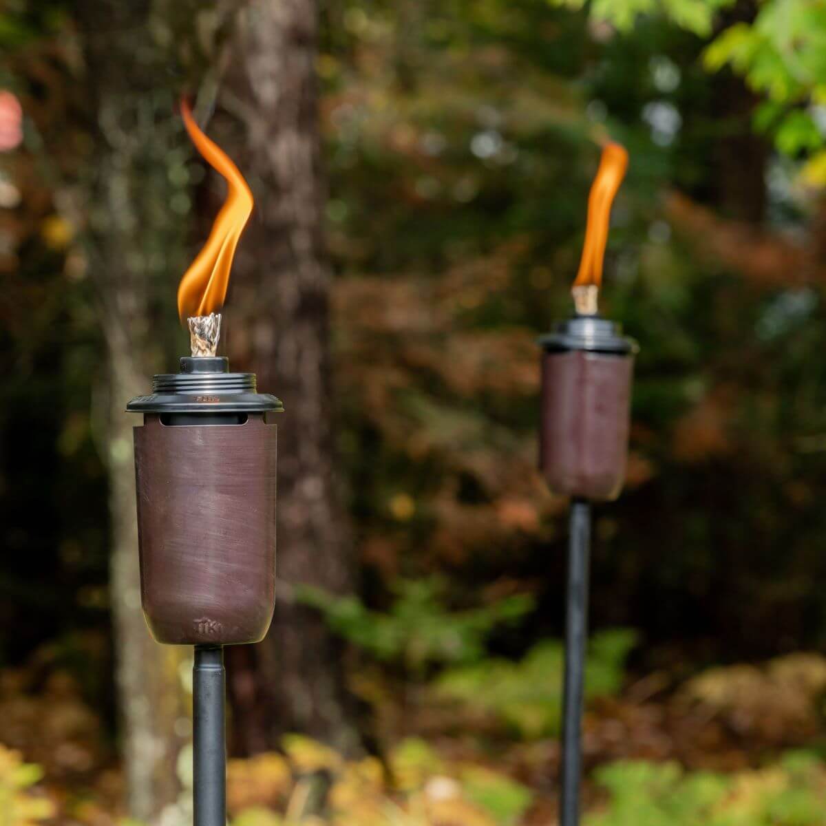 Two brown TIKI® torches with bright flames, set outdoors against a background of trees and greenery.