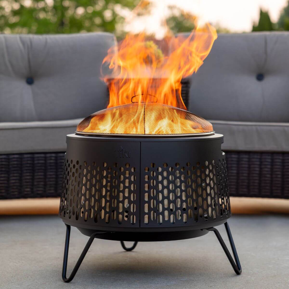 TIKI® fire pit with mesh cover, burning brightly on a patio in front of an outdoor sofa.