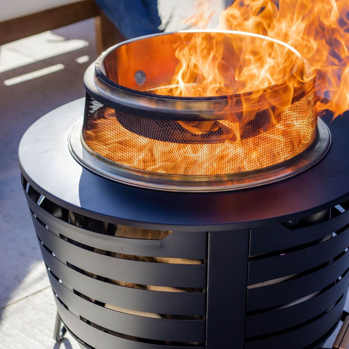 A TIKI® patio fire pit with a bright flame, featuring a black metal base and mesh screen for outdoor use.