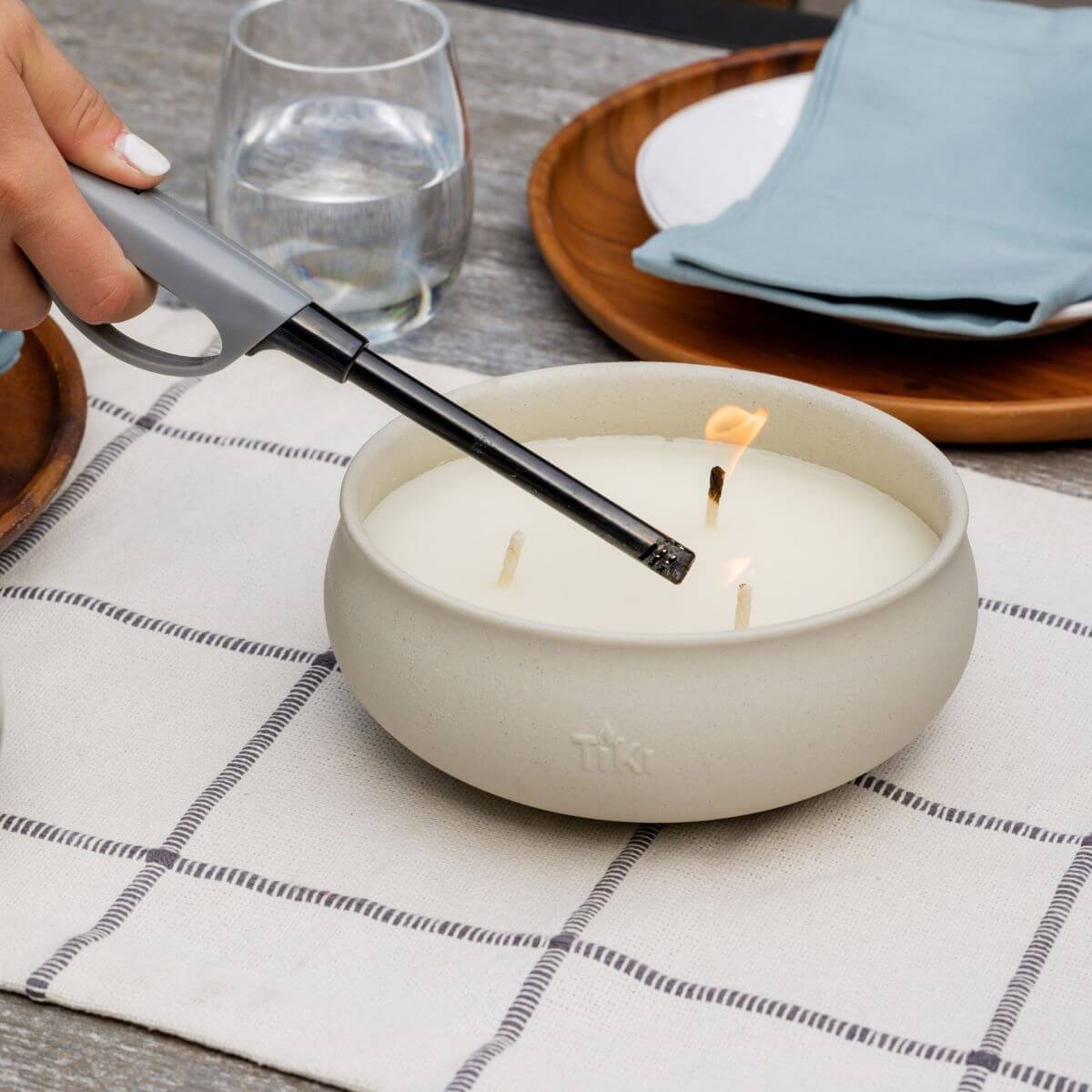 Hand lighting a TIKI citronella candle bowl on a table set for outdoor dining.