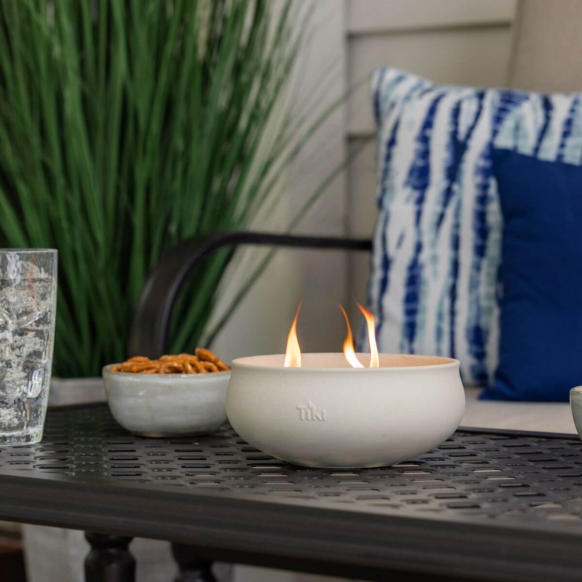 TIKI tabletop fire bowl with flames on a patio table, next to snacks and a glass, outdoor seating in background.