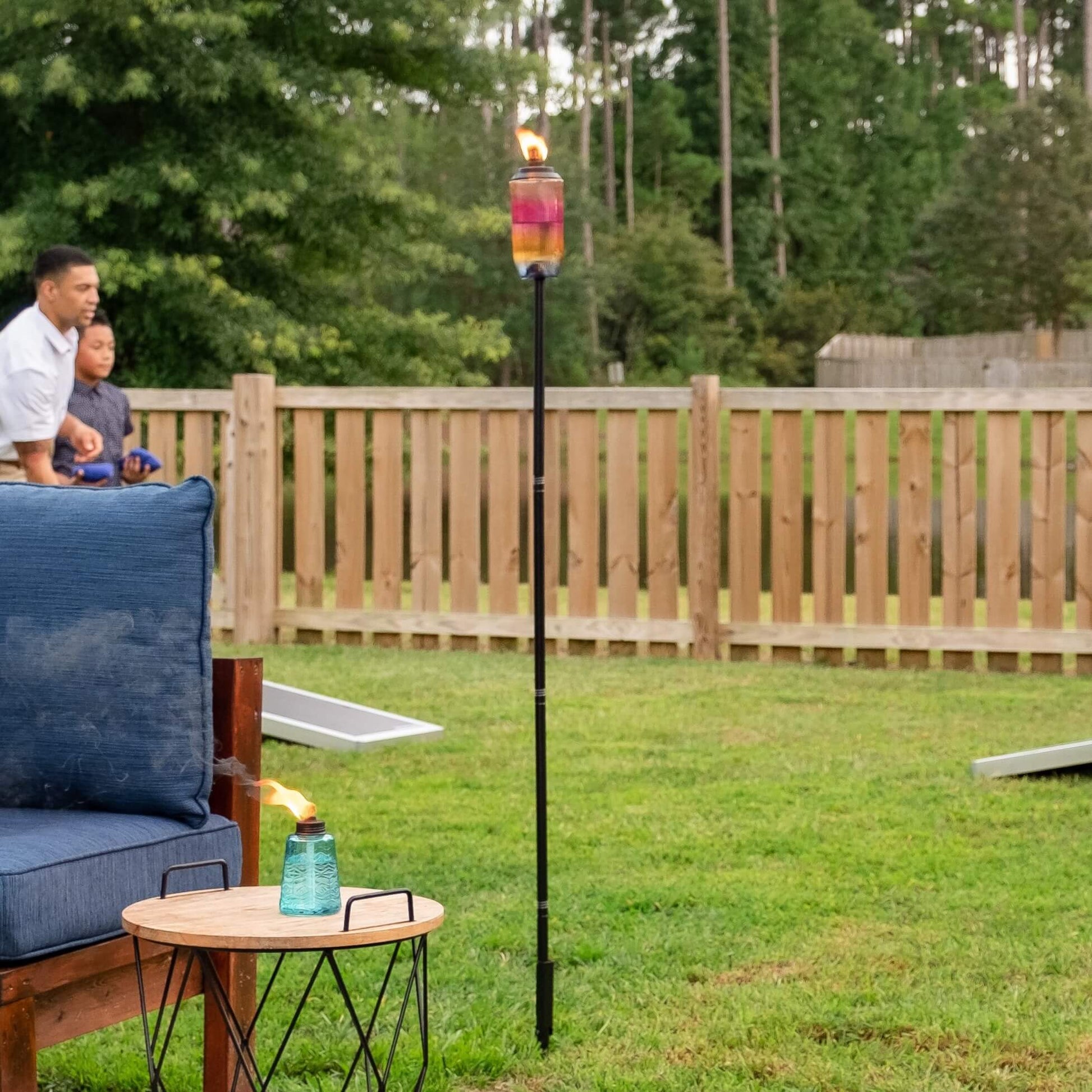 Colorful TIKI® Brand torch and tabletop lantern add ambiance to a backyard gathering with games and outdoor seating.