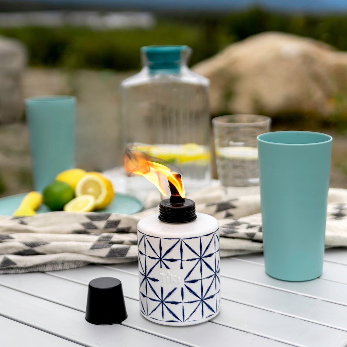 TIKI ceramic table torch with geometric pattern, lit on an outdoor table with drinks and sliced lemons in the background.