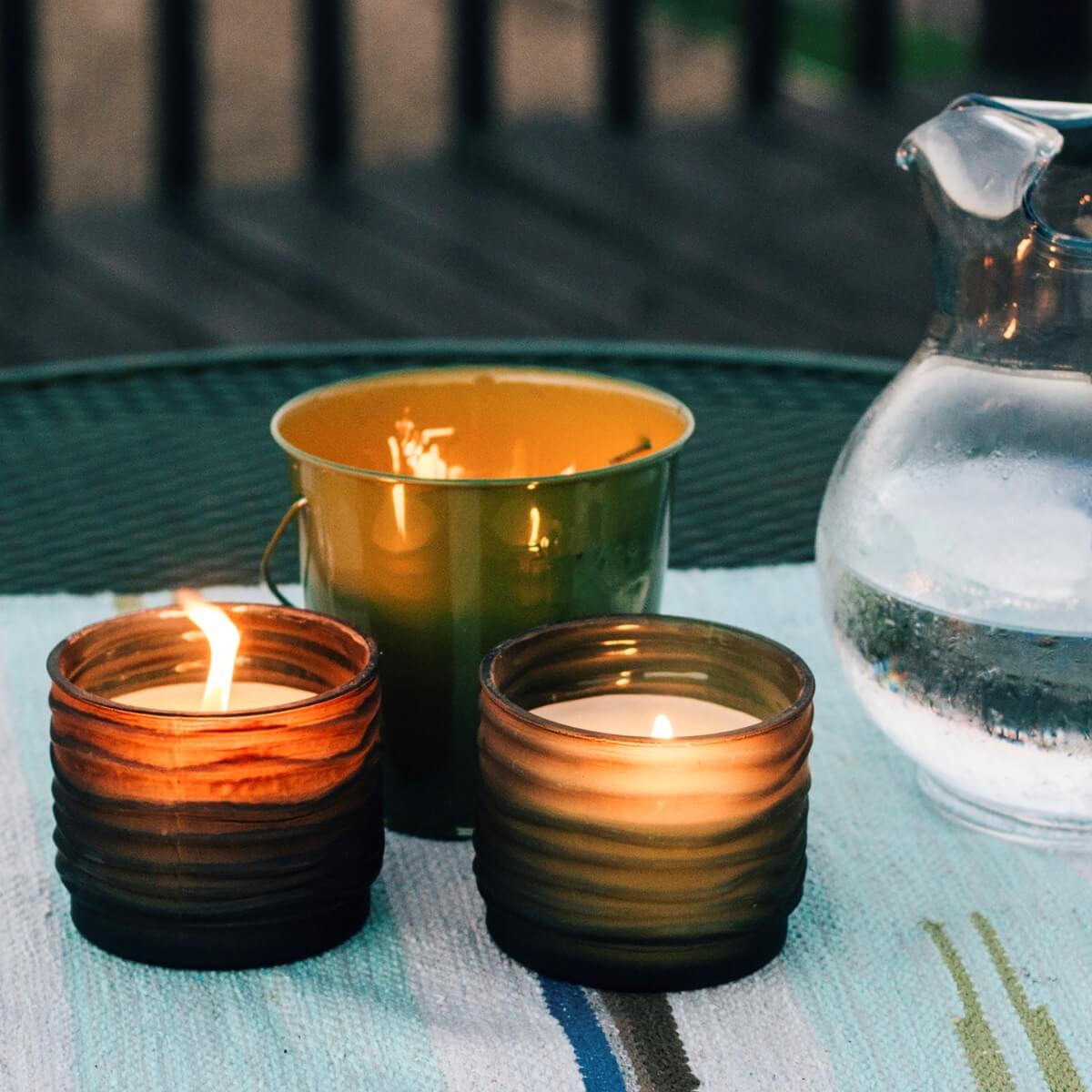 Two TIKI® Brand citronella candles and a green bucket candle on an outdoor table, next to a glass pitcher of water.
