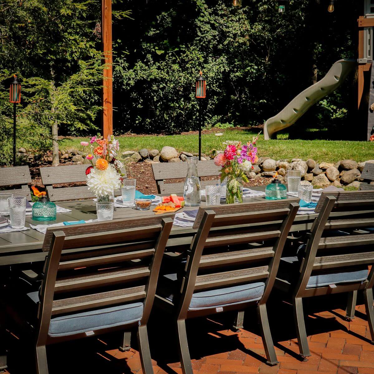 Outdoor dining table with flowers, glassware, and TIKI torches in a backyard setting; playground slide in background.
