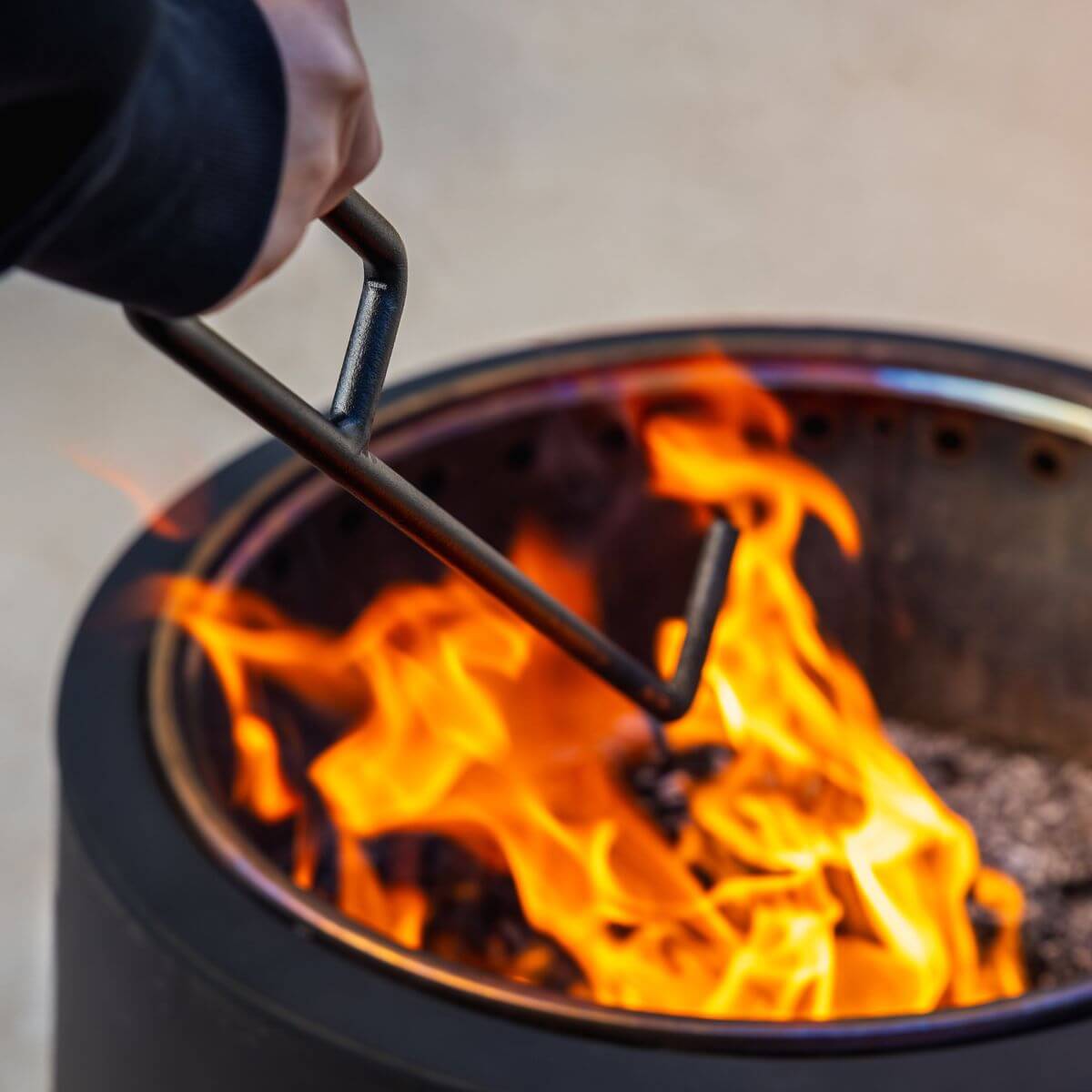 Hand using a fire poker to adjust burning wood in a TIKI® fire pit. Bright flames and metal rim are visible.
