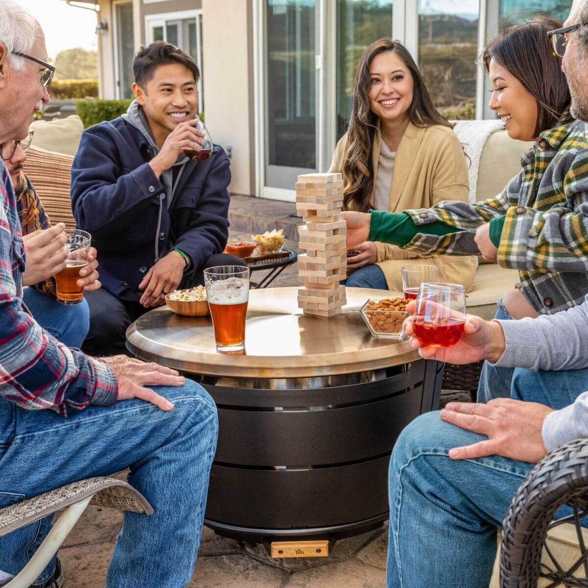 Group enjoying drinks, snacks, and a game of Jenga around a TIKI® fire pit table on a patio.
