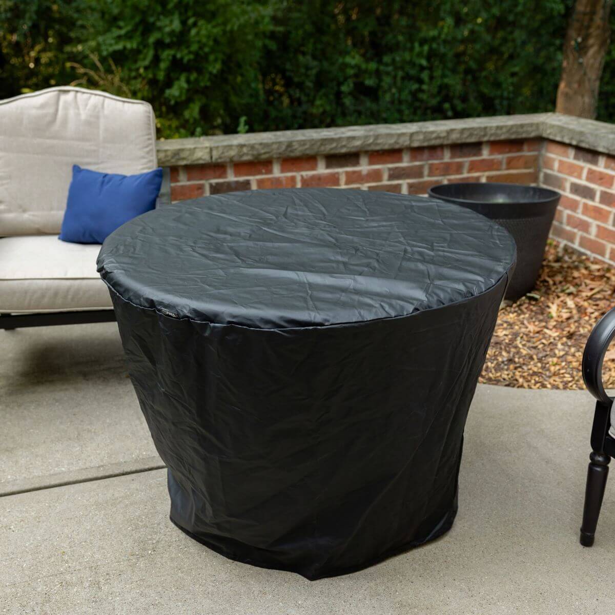 Black protective cover on a round outdoor fire pit, placed on a patio near seating and greenery.