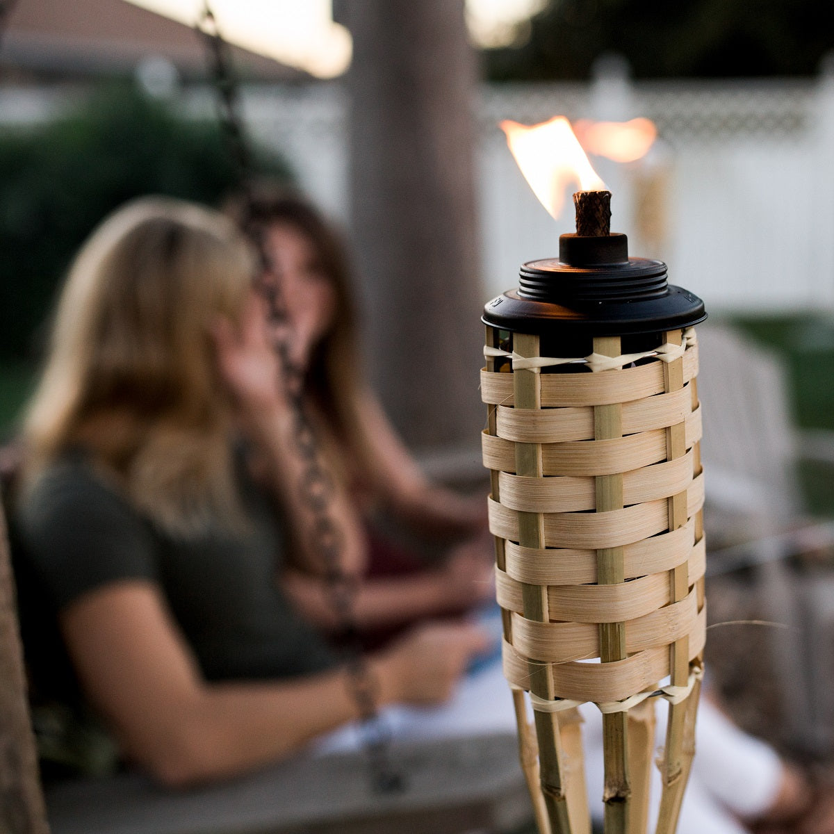Lit bamboo TIKI® torch in foreground; two women sit and talk outdoors in a backyard setting.