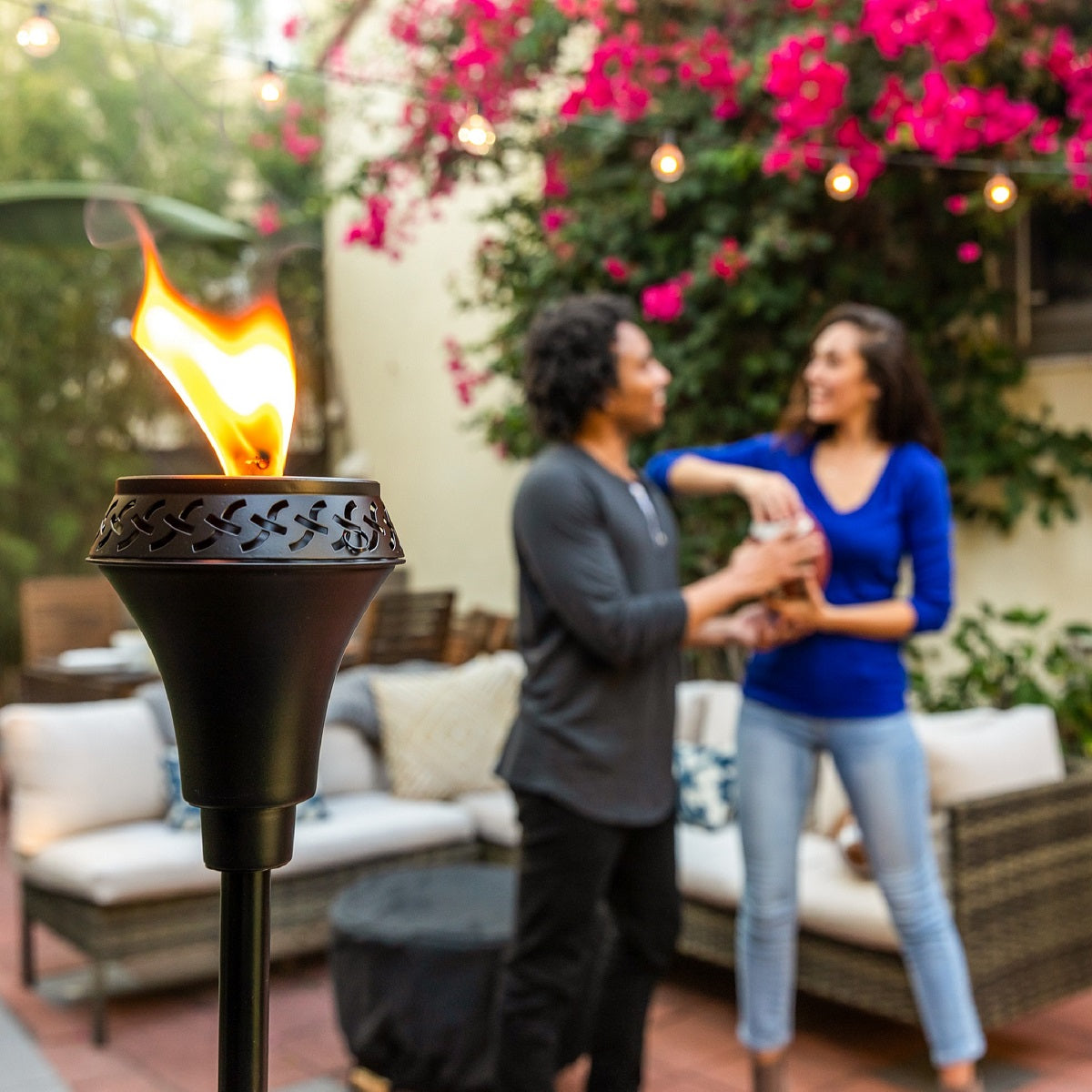 Lit TIKI® Brand torch in a cozy patio setting with two people enjoying an outdoor gathering under string lights and flowers.