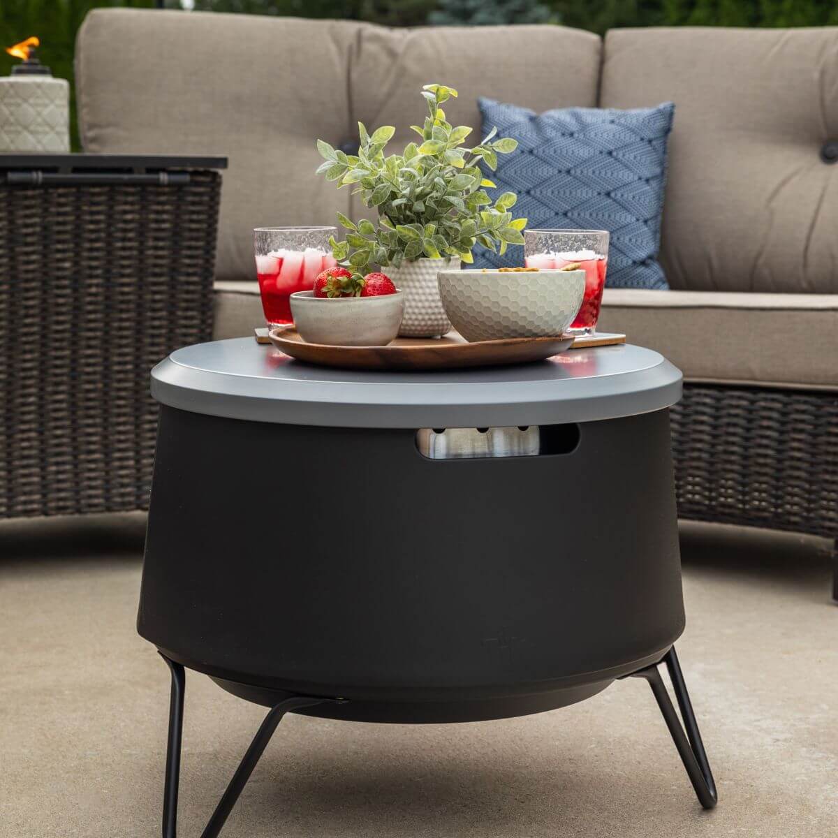 Round black patio table with drinks, snacks, and a plant on top, set in front of an outdoor wicker sofa.