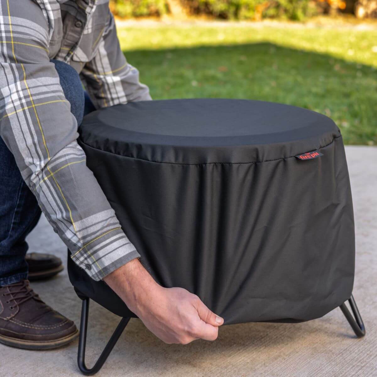 Person placing a black protective cover over a TIKI® fire pit on a patio, with grass in the background.