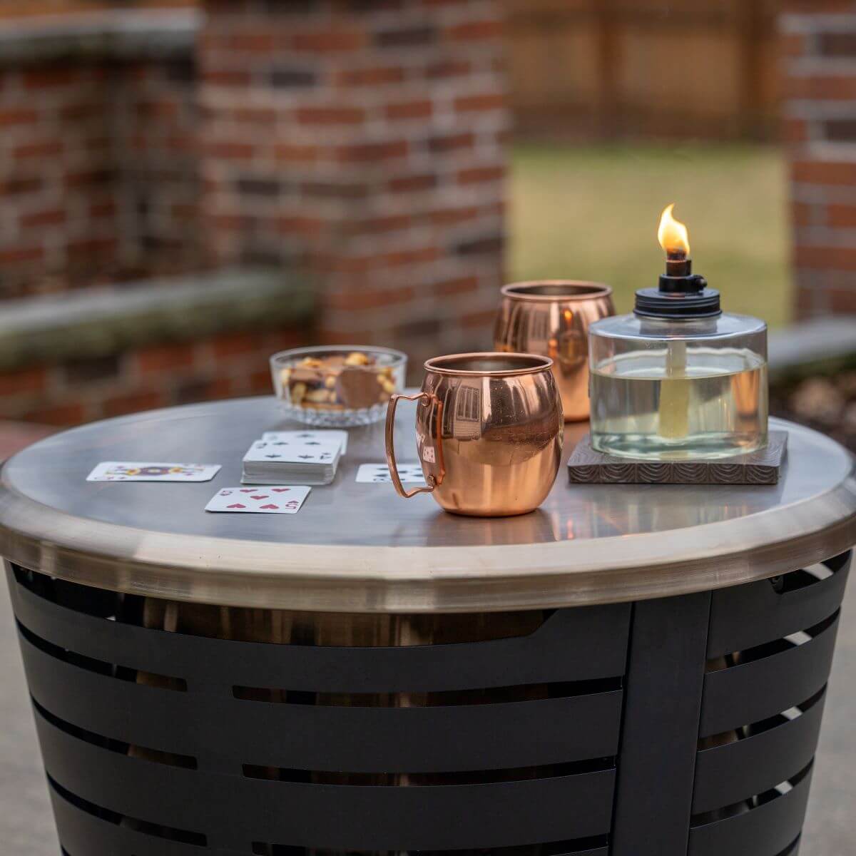 Outdoor table with TIKI® torch, copper mugs, playing cards, and snacks, set for a cozy backyard gathering.