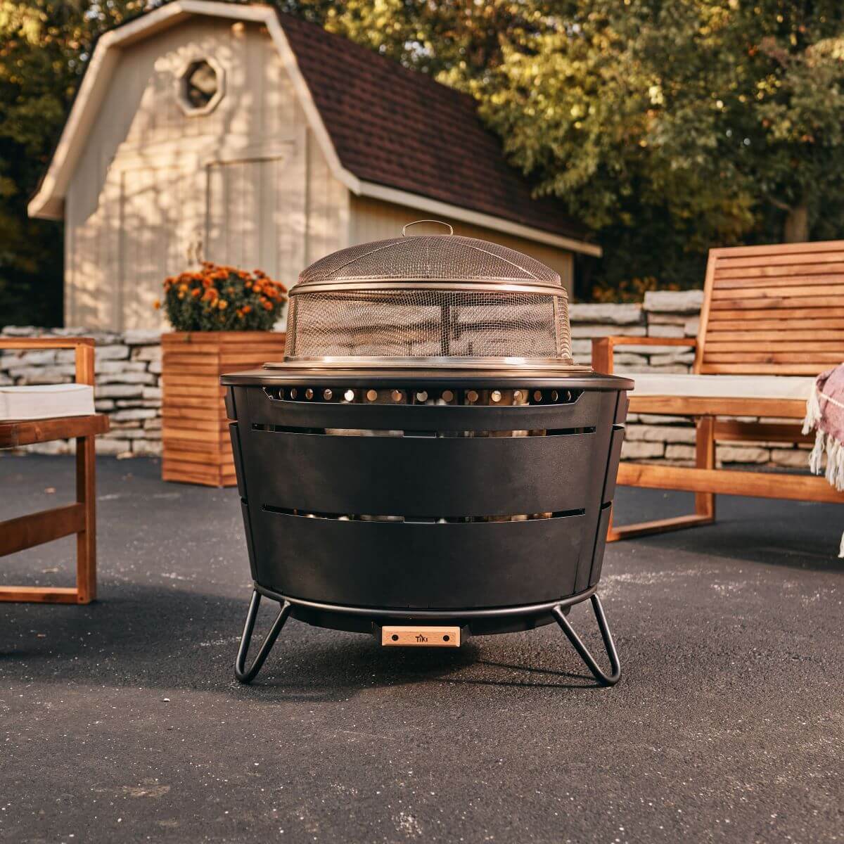 Black TIKI fire pit with mesh cover on patio, surrounded by wooden outdoor chairs; shed and plants in background.