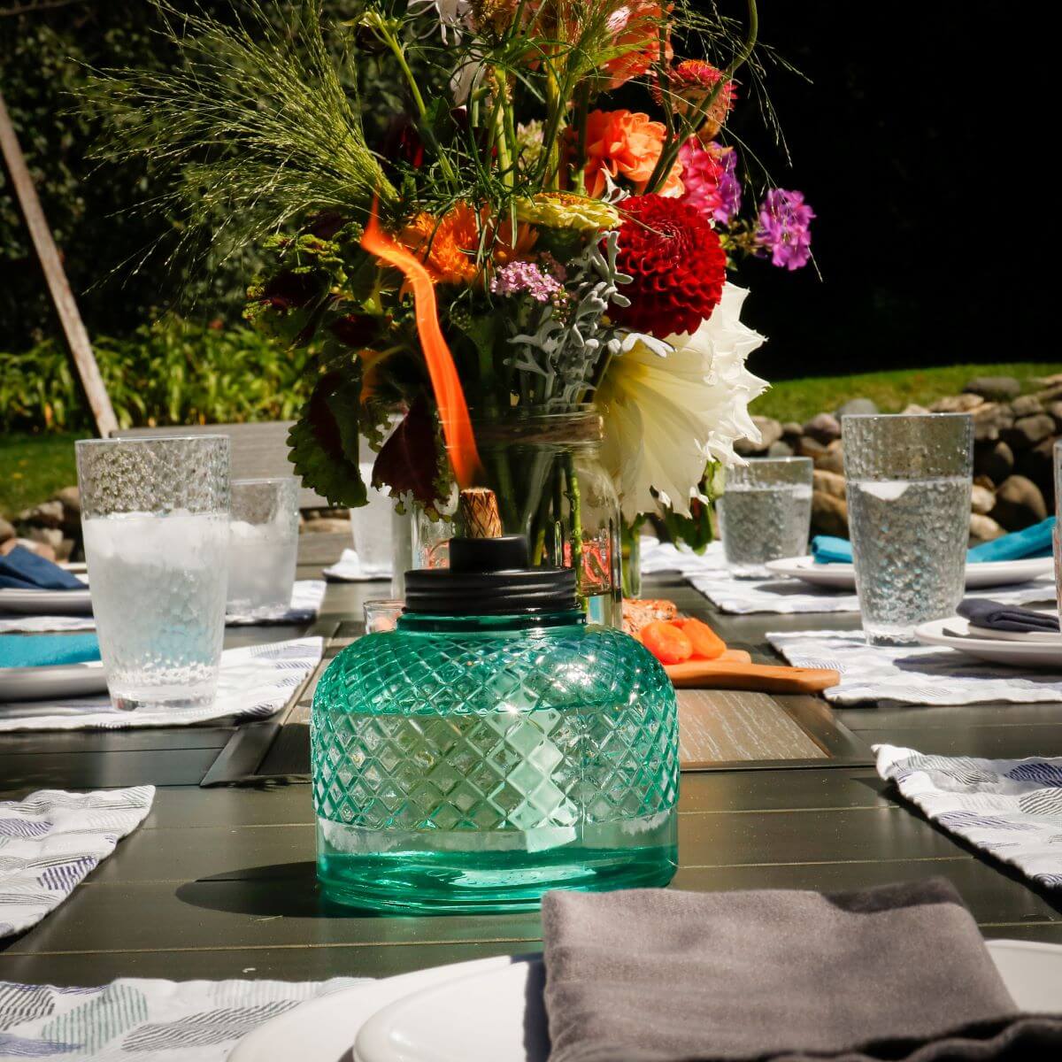 A teal TIKI® Brand table torch with a diamond pattern sits on an outdoor dining table set for a gathering.