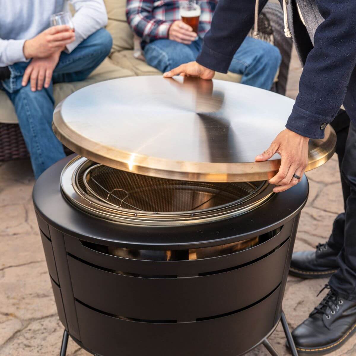 Person lifting lid off a TIKI Brand fire pit on a patio, with people sitting and holding drinks in the background.