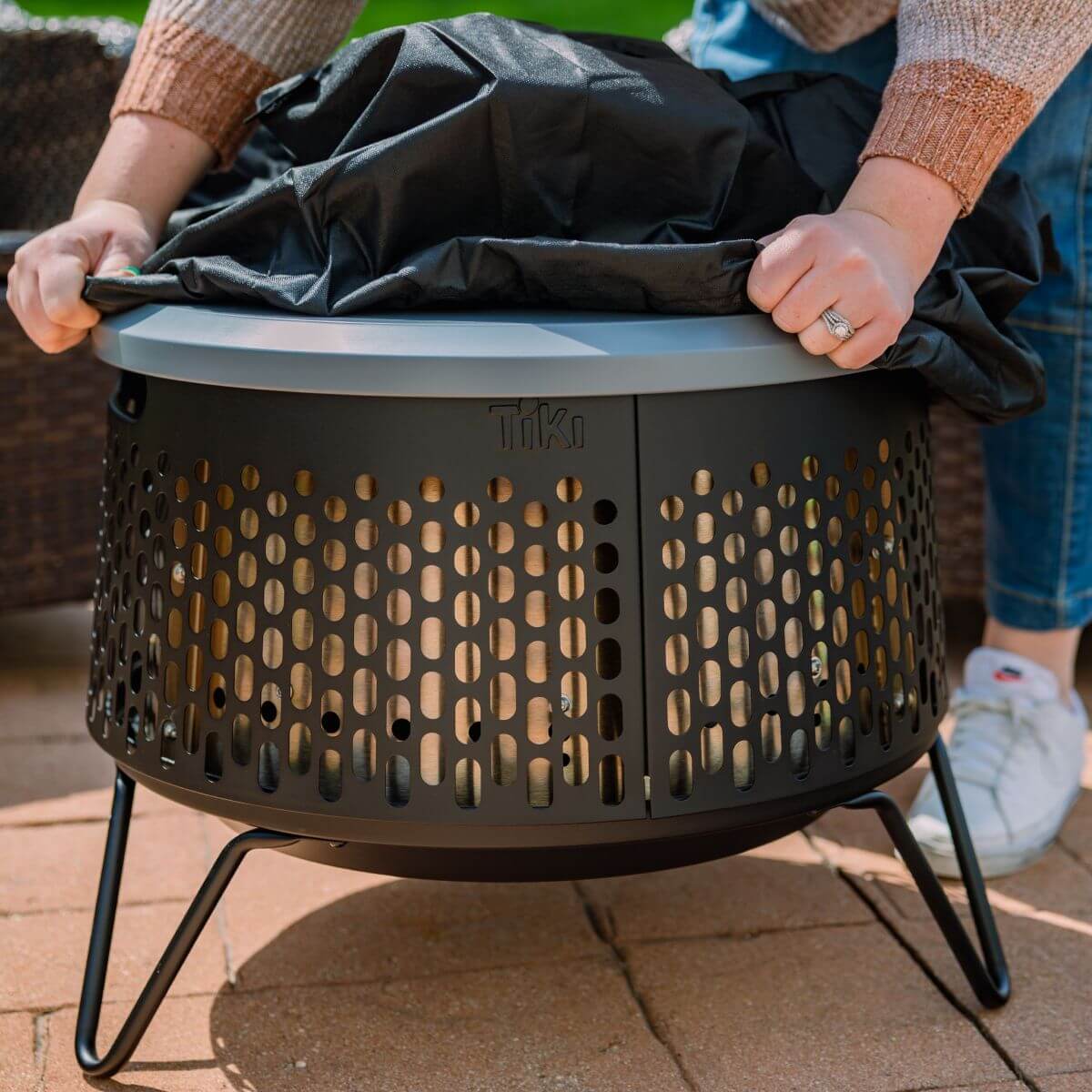 Person placing a weatherproof cover on a TIKI fire pit outdoors; easy protection for patio use.