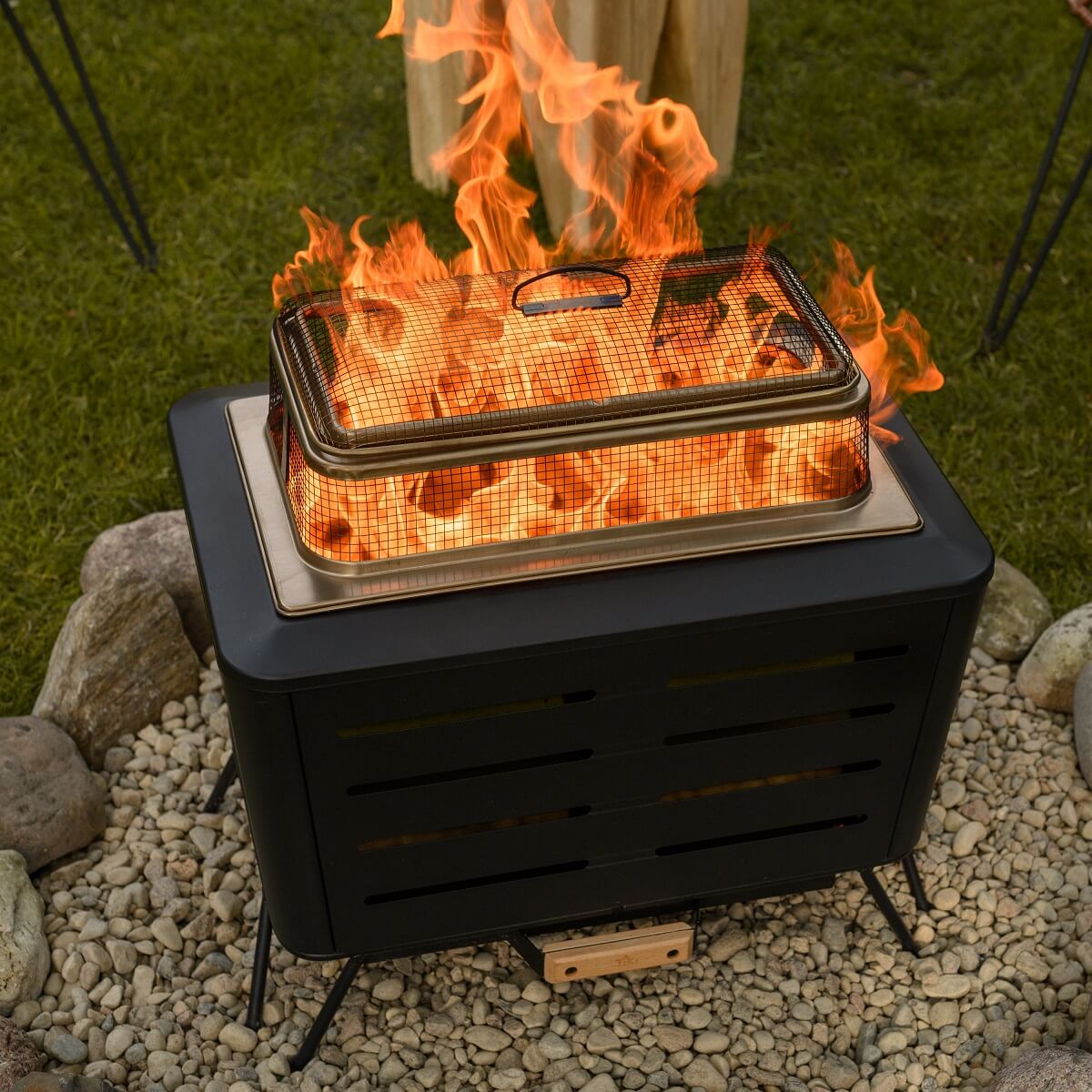 Rectangular TIKI fire pit with a mesh spark screen, burning brightly on a stone patio in a grassy backyard.