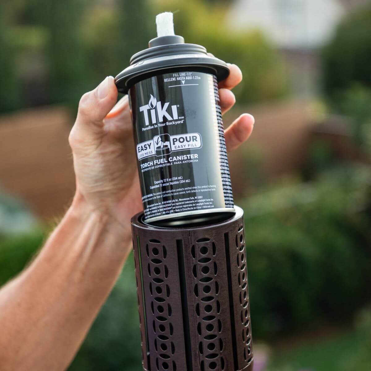 Hand holding a TIKI torch fuel canister above a decorative torch holder outdoors, ready for easy installation.