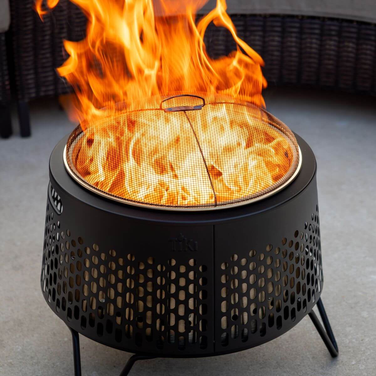 Black TIKI fire pit with mesh screen cover, showing bright flames, on a patio surface.