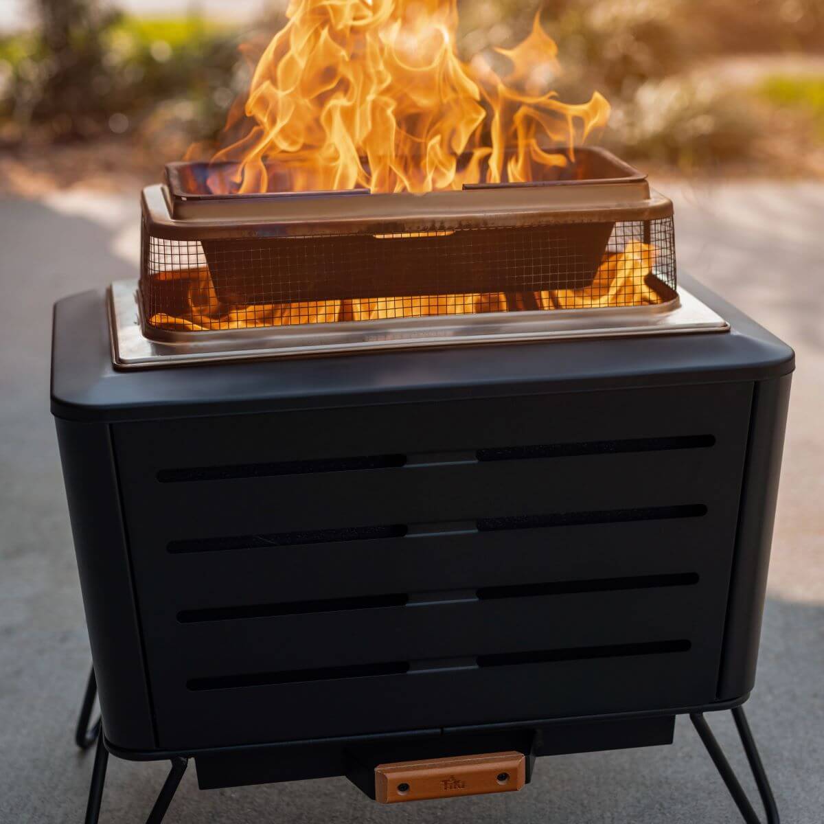 Black TIKI fire pit with mesh screen, burning bright flames outdoors on a patio.