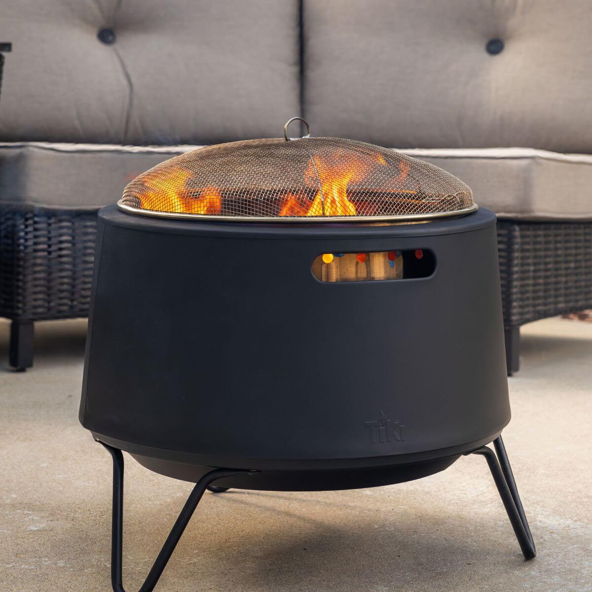 Black TIKI fire pit with mesh cover, burning firewood, placed on a patio near outdoor seating.