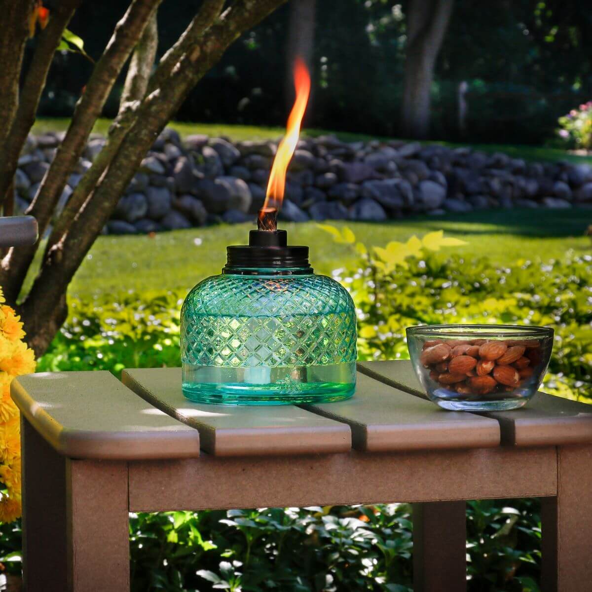 A turquoise TIKI® glass table torch with a flame, next to a bowl of almonds, on a patio table outdoors.
