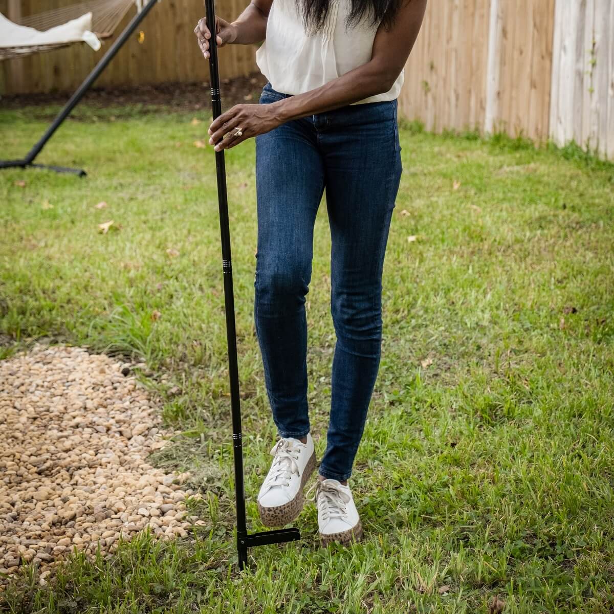 Woman installing a TIKI torch stake in backyard grass, using foot to press stake into ground.