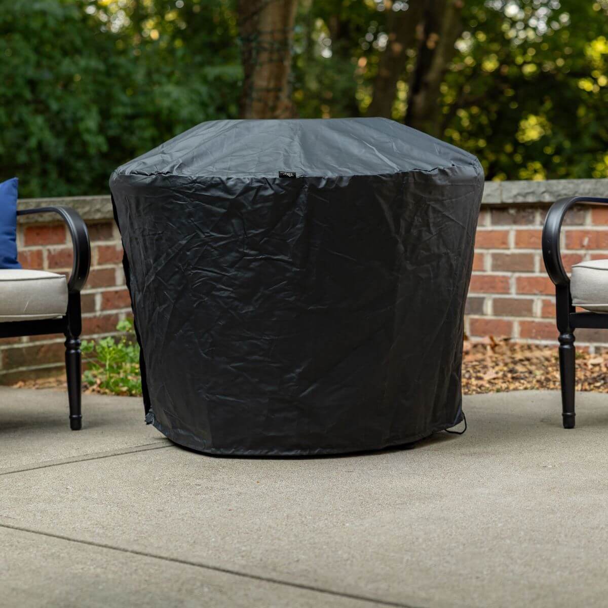 Black protective cover over a TIKI fire pit on a patio, with two chairs and greenery in the background.