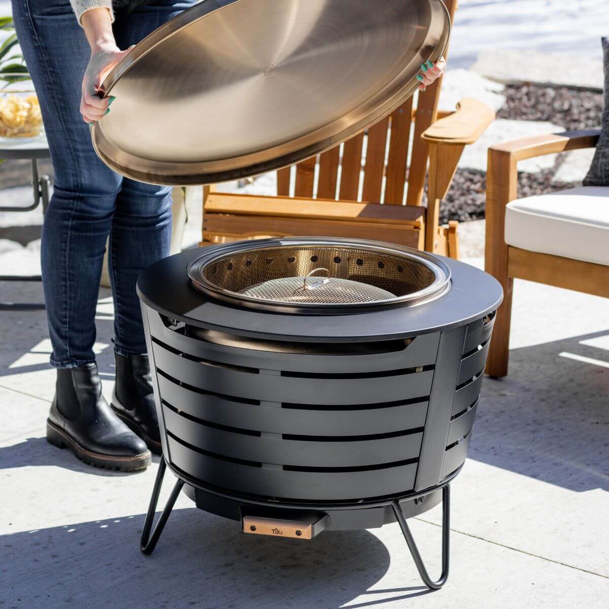 Person lifting lid off a TIKI Brand fire pit on a patio, surrounded by outdoor seating.