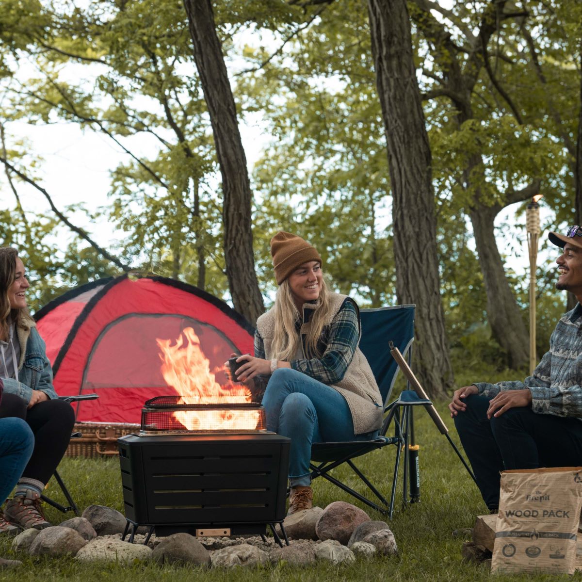 Three people enjoy a campfire in a TIKI® fire pit near a red tent, surrounded by trees and TIKI® Wood Pack boxes.