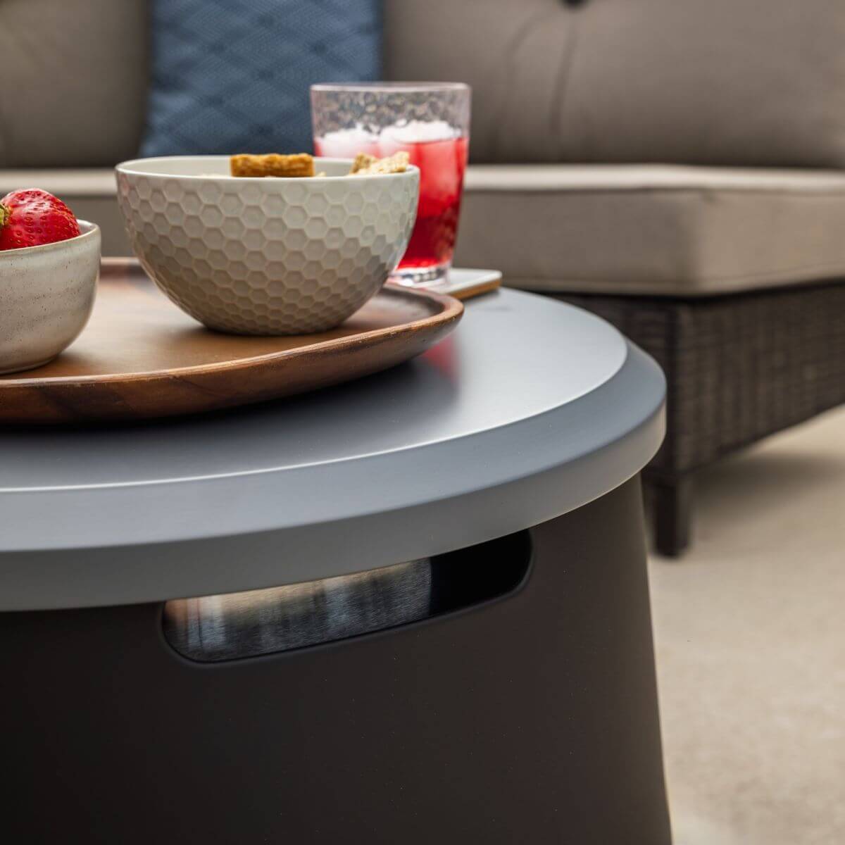 Outdoor patio table with snacks and drinks on top, next to a cushioned sofa. Accessible, modern design from TIKI Brand.