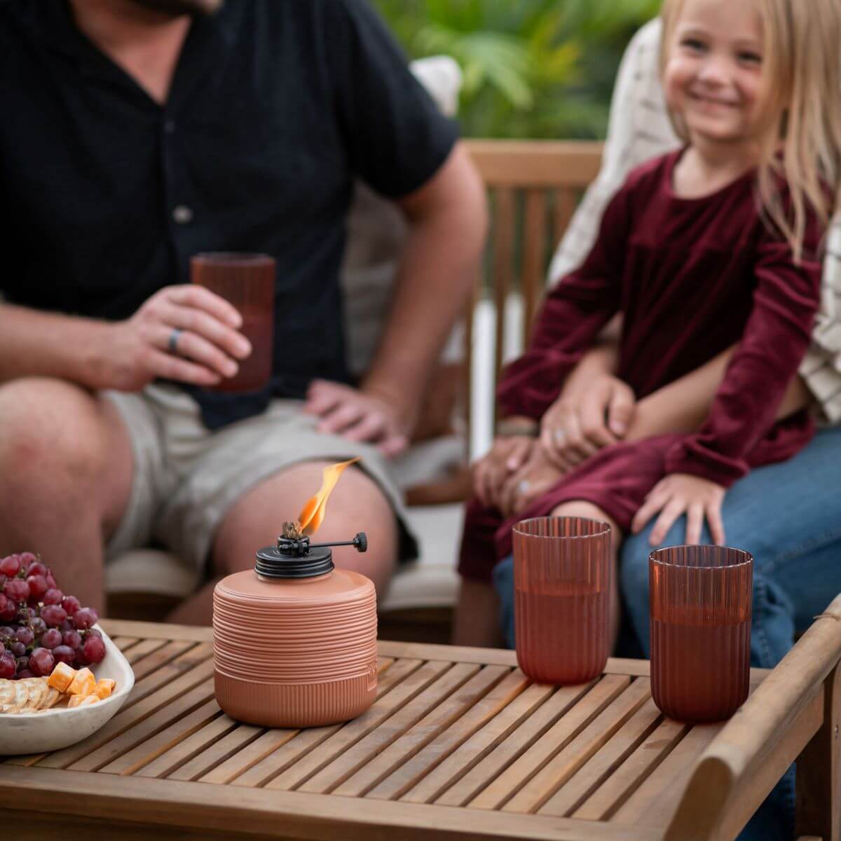 Family enjoys outdoor seating with a TIKI® tabletop fire pit, drinks, and snacks on a wooden patio table.