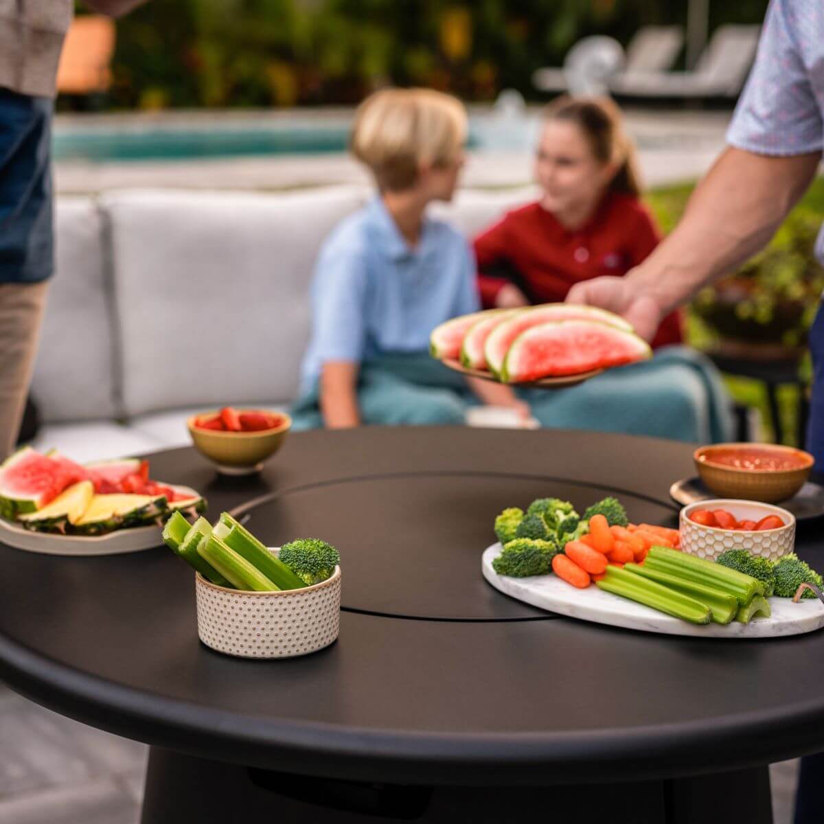 Outdoor table with fresh fruit, veggies, and dip; people relaxing by a pool in a backyard setting.