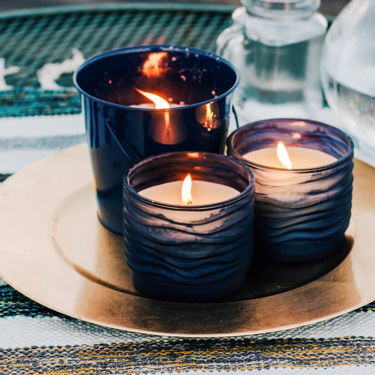 Three lit TIKI® Brand candles in blue glass and metal holders on a gold tray, set on an outdoor table.