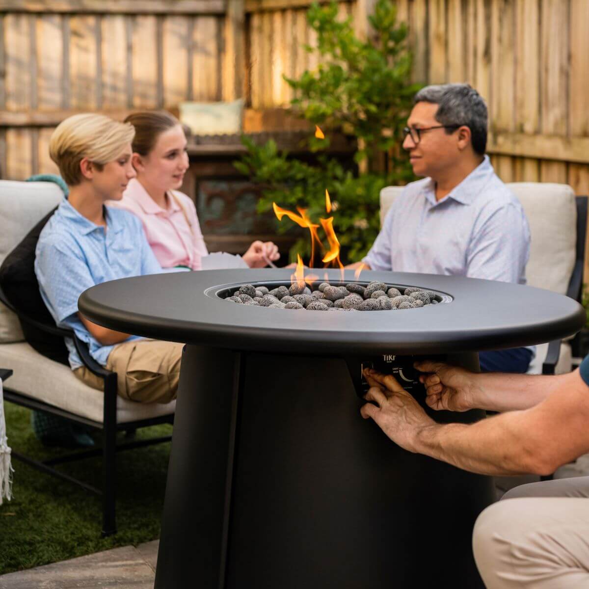 People enjoy a backyard gathering around a TIKI Brand fire pit table with adjustable flame and modern design.