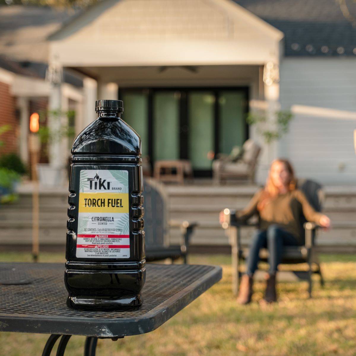 TIKI Brand Citronella Torch Fuel bottle on a patio table in a backyard, with a house and outdoor seating in the background.
