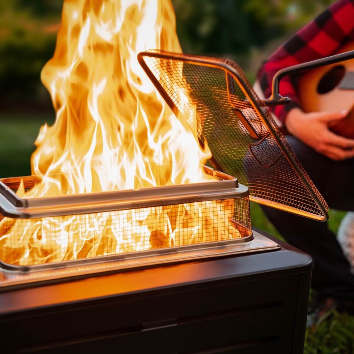 A TIKI® fire pit with a mesh screen cover, burning brightly outdoors. Person in background playing guitar.