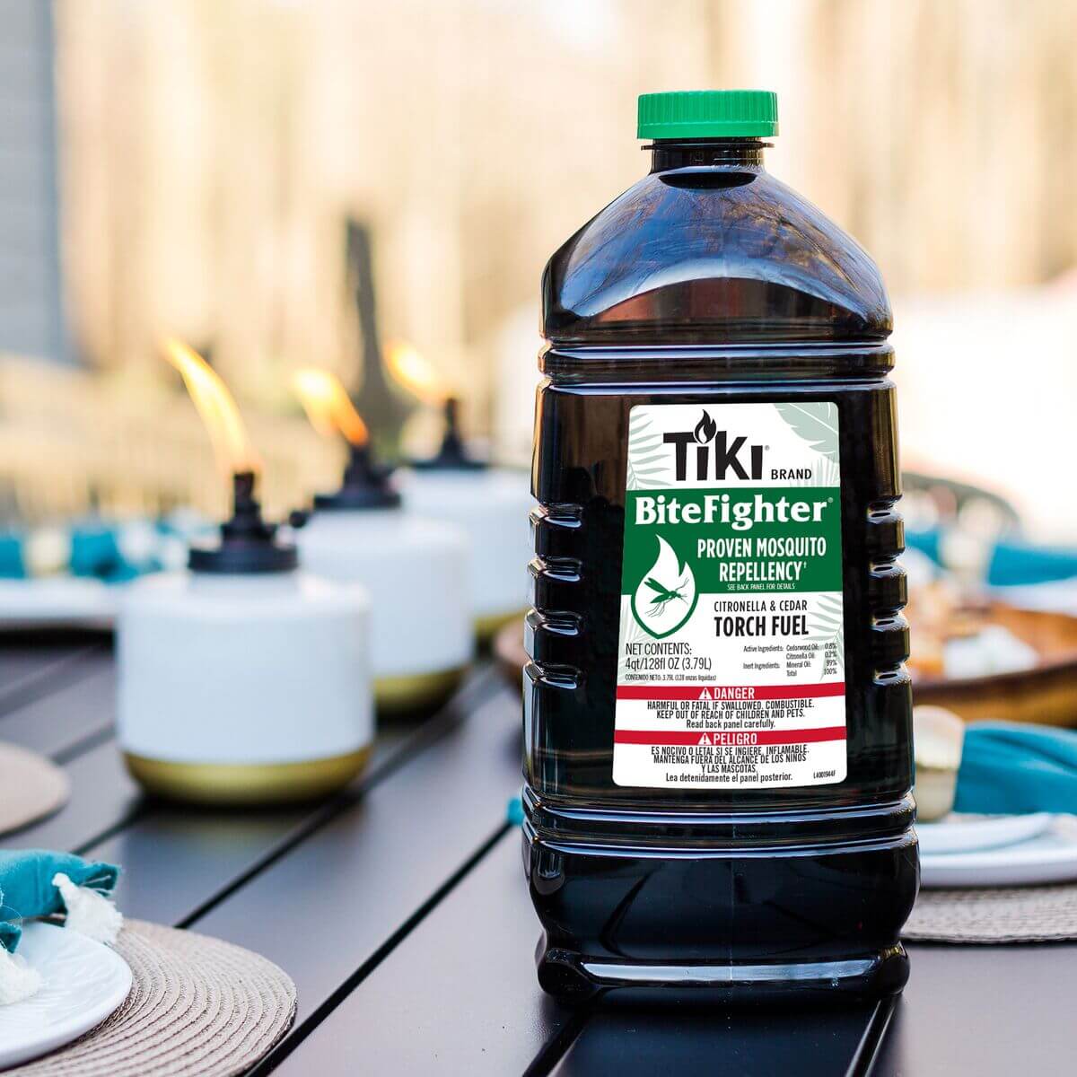 TIKI Brand BiteFighter Torch Fuel bottle on outdoor table; torches lit in background for mosquito repellency.