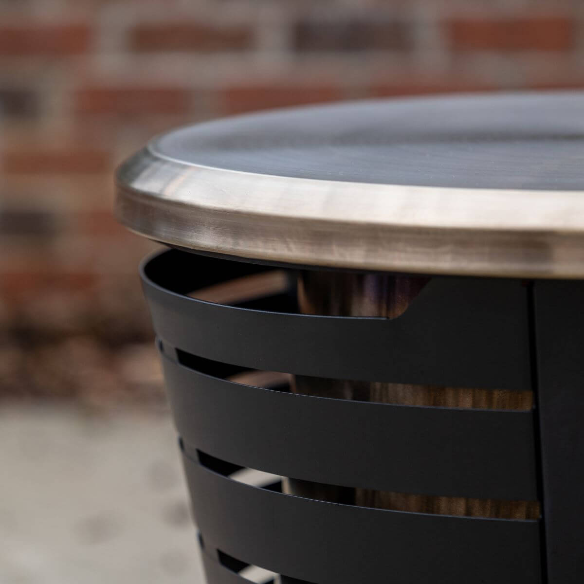 Close-up of a TIKI® fire pit rim and black metal slatted side, with a brick wall in the background.