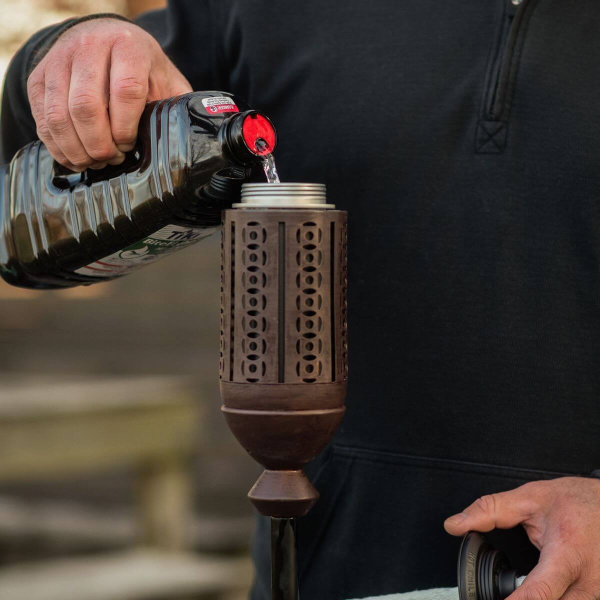 A person pours TIKI Brand torch fuel into a brown outdoor torch, preparing it for use.
