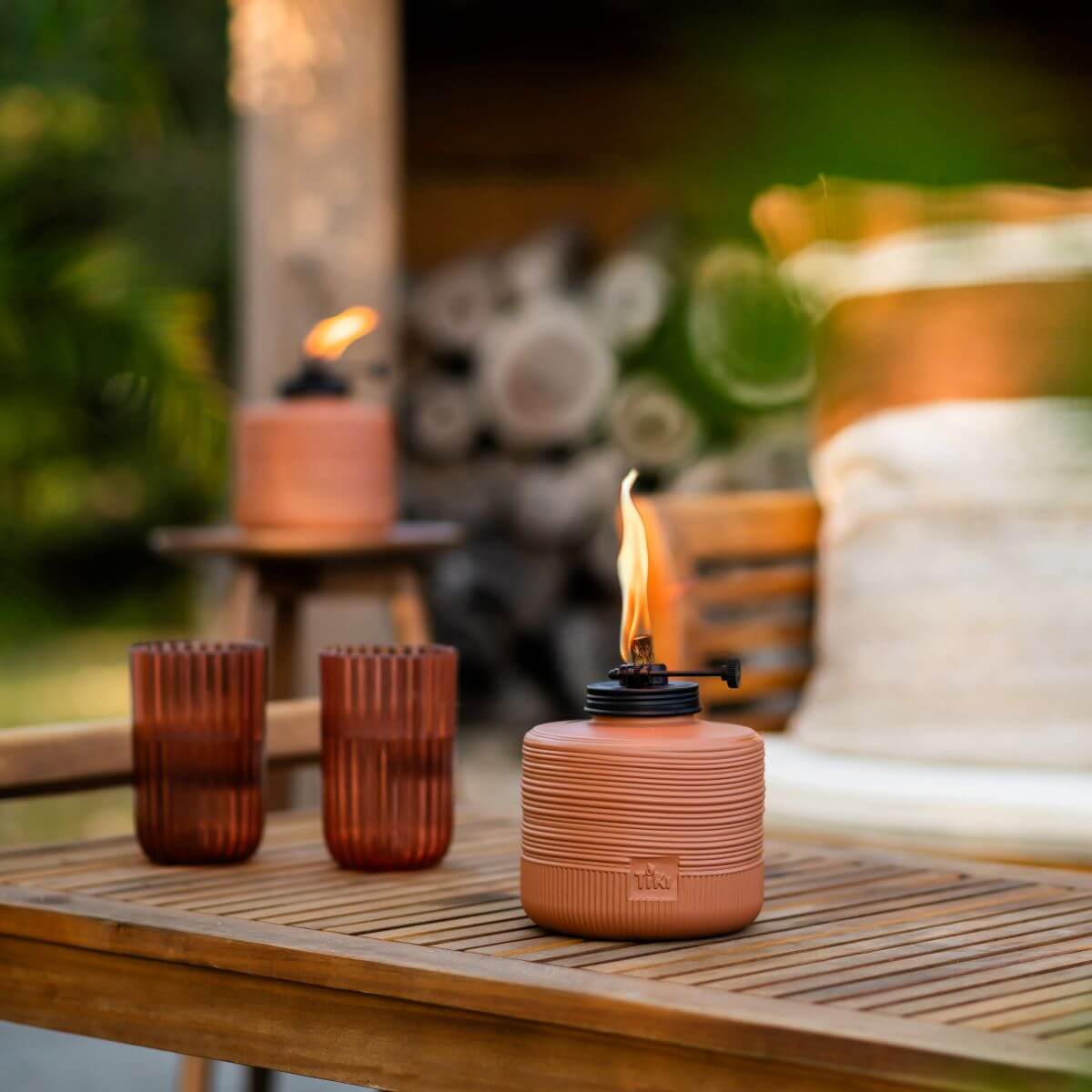 Terracotta TIKI® tabletop torch with a bright flame on a wooden patio table, next to two ribbed brown glasses outdoors.