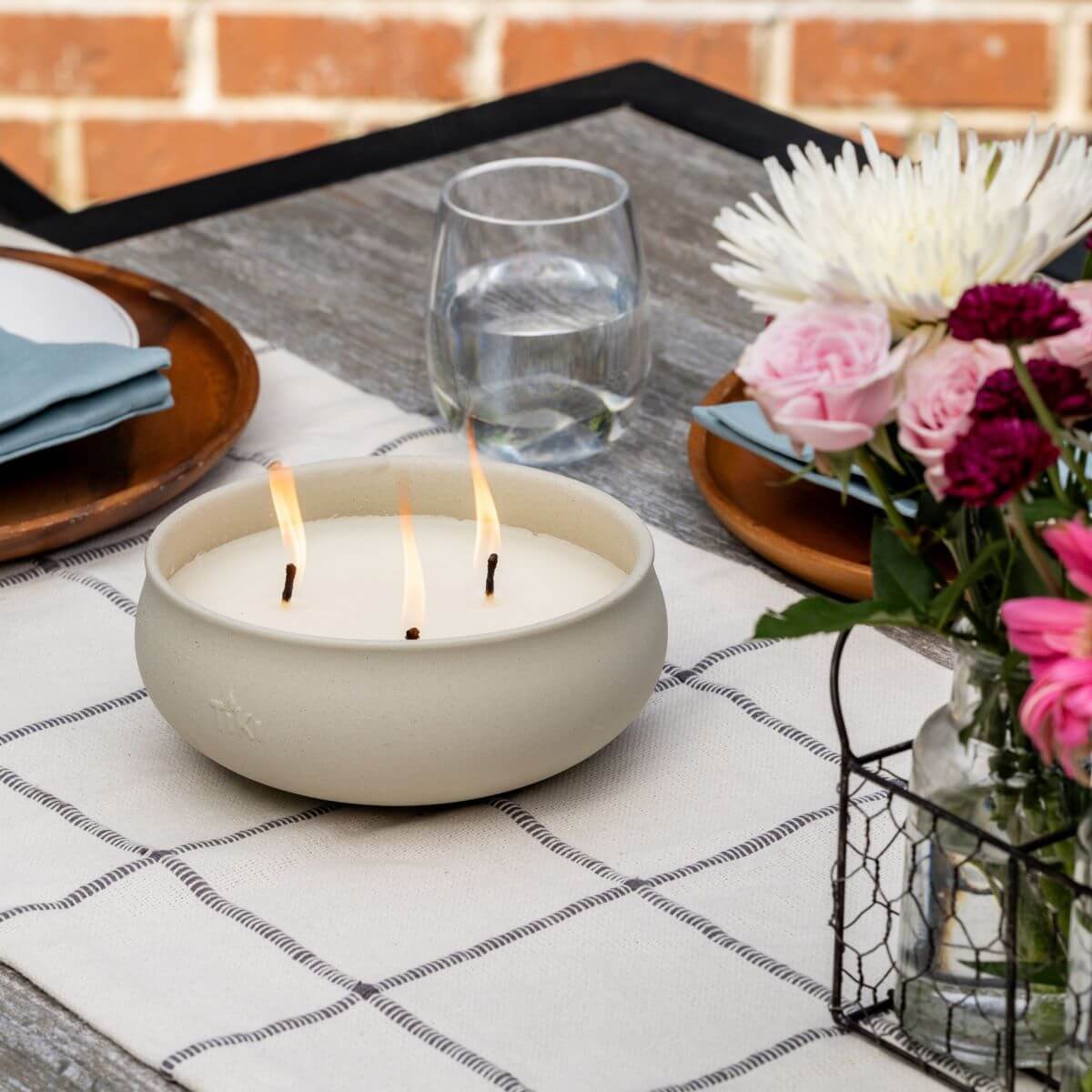 Three-wick TIKI® Brand tabletop citronella candle burning on an outdoor dining table set with flowers and place settings.