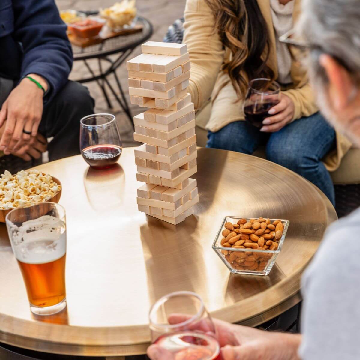 Friends play Jenga outdoors on a round table with drinks, popcorn, and almonds, enjoying a casual gathering.