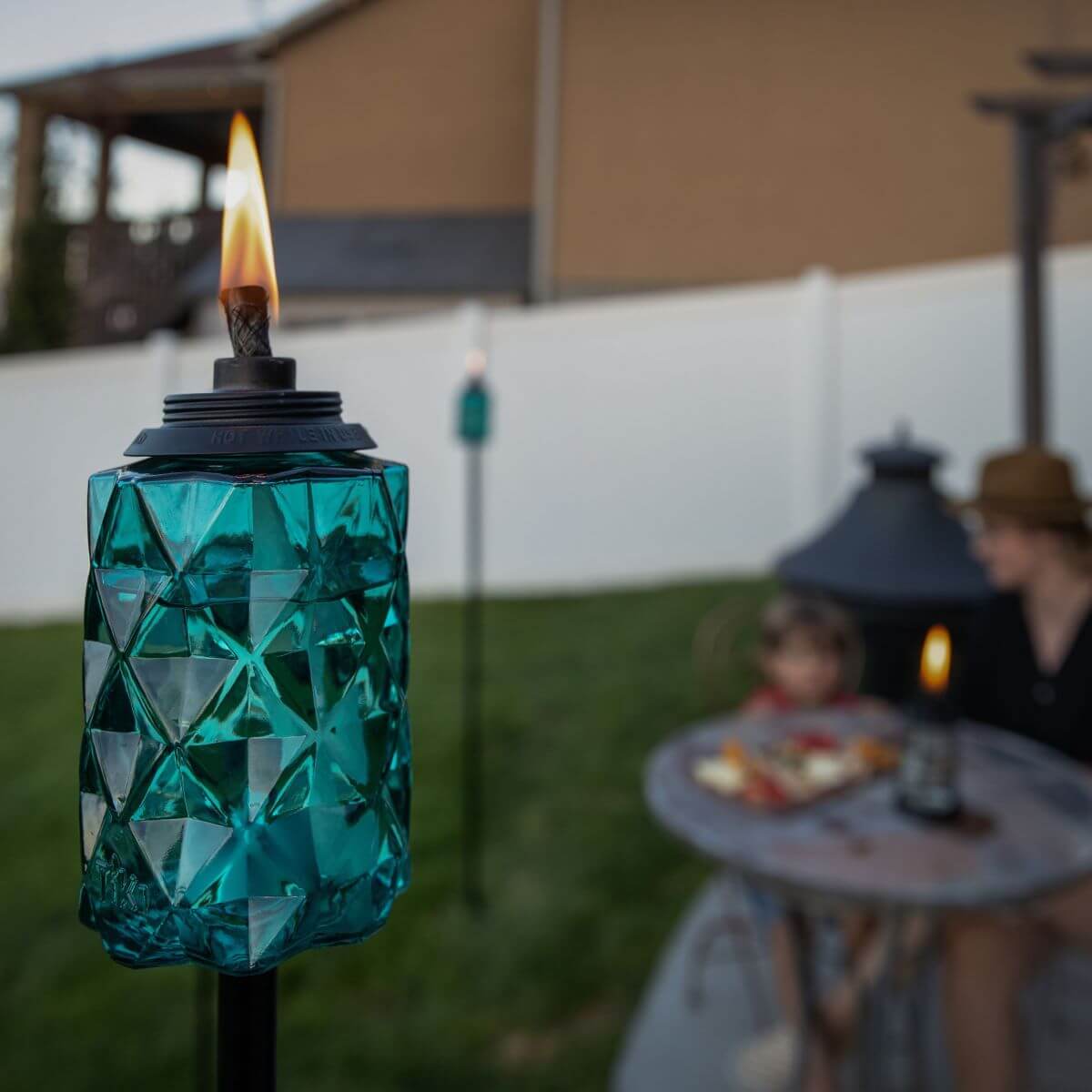 A turquoise TIKI® Brand torch burns in a backyard, adding ambiance to an outdoor seating area with a table and snacks.