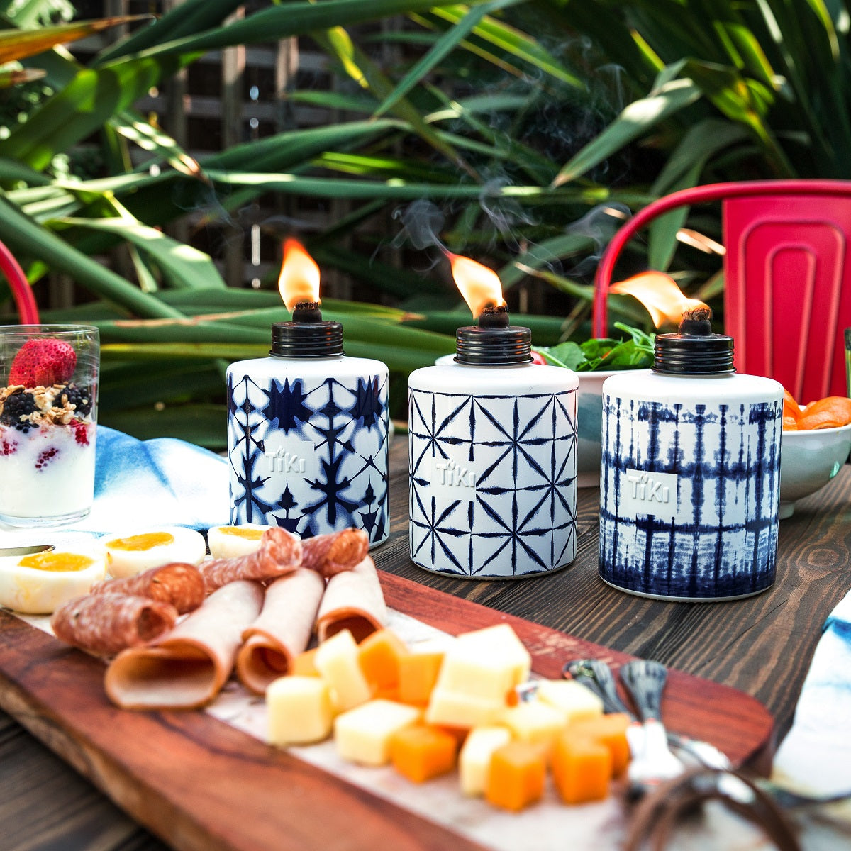 Three patterned TIKI table torches with flames on an outdoor table, surrounded by snacks and greenery.