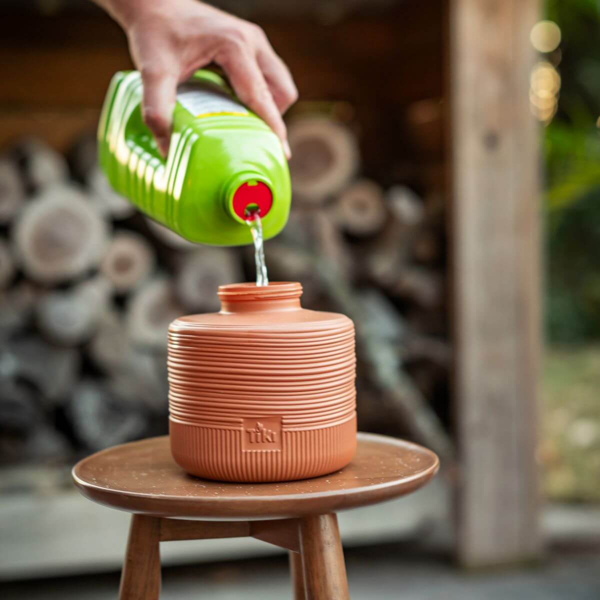 A hand pours TIKI-brand torch fuel from a green bottle into a terracotta torch canister outdoors.