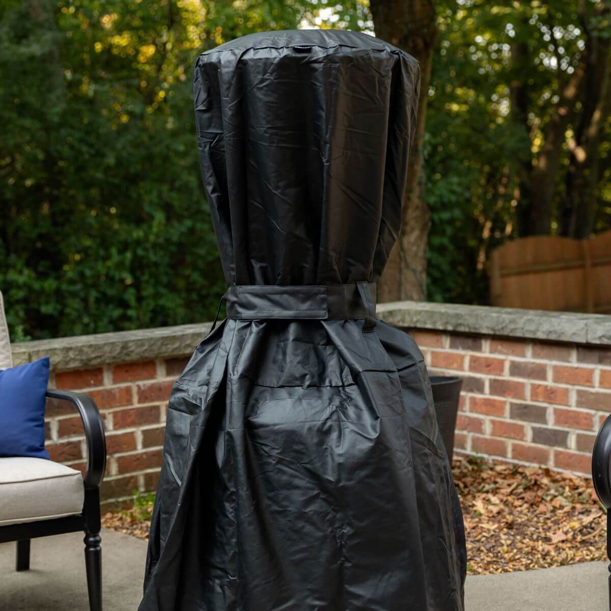 Black outdoor fire pit cover protects TIKI fire pit from weather; shown on patio with trees and brick wall in background.